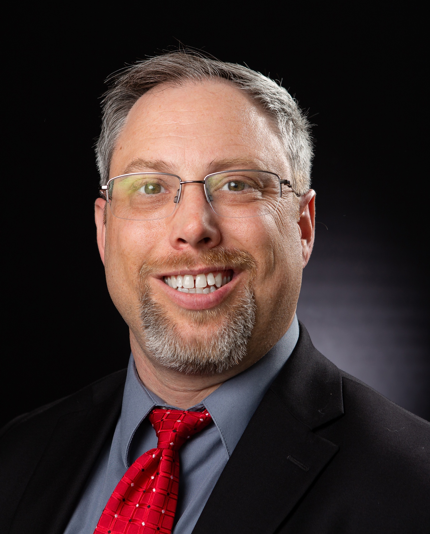 Connor in a photo studio wearing a black blazer, blue shirt, and red tie smiling at the camera
