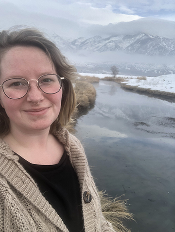 Selfie style image of Megan by a mountain lake covered in snow.