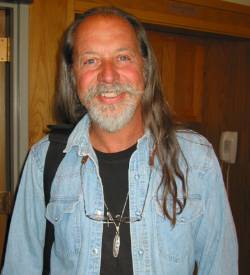 Stephen standing outstand of an office. He is smiling at the camera with reading classes around his neck, a black t-shirt, and light blue denim shirt.